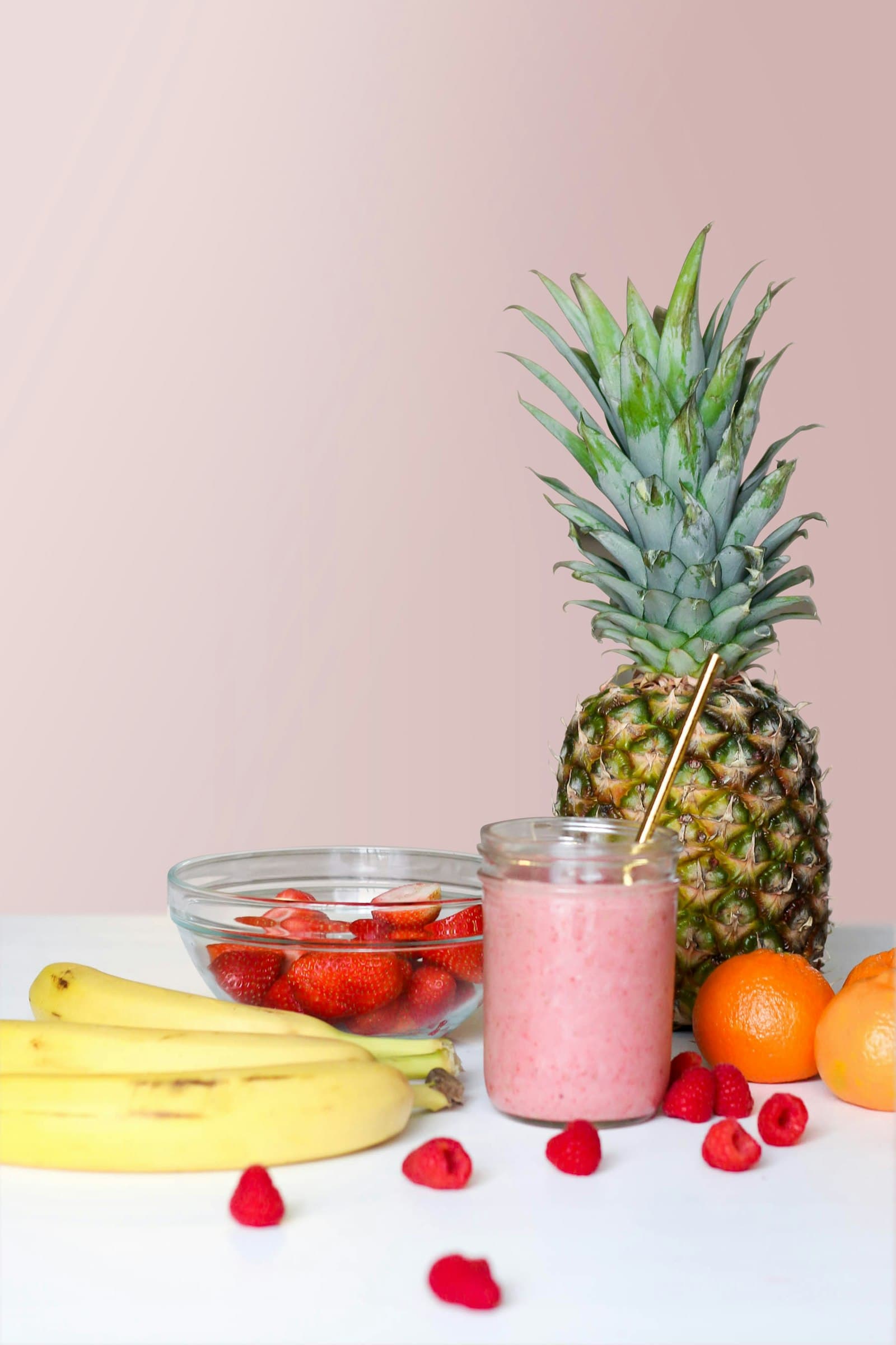 A pink smoothie surrounded by bananas, strawberries, oranges and a pineapple