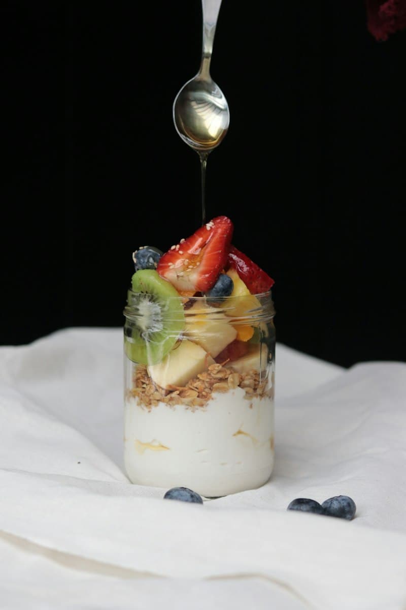 Honey drizzling from a spoon onto a jar of yoghurt, granola, kiwi and strawberry
